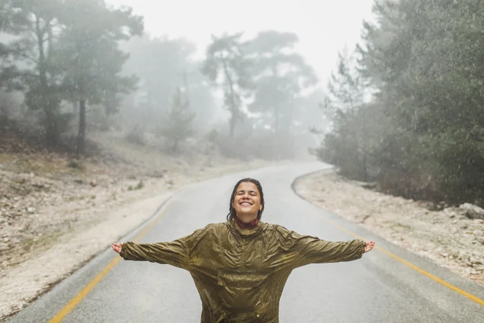 Woman smiling with arms outstretched standing in the rain on a misty forest road, representing freedom and wellbeing