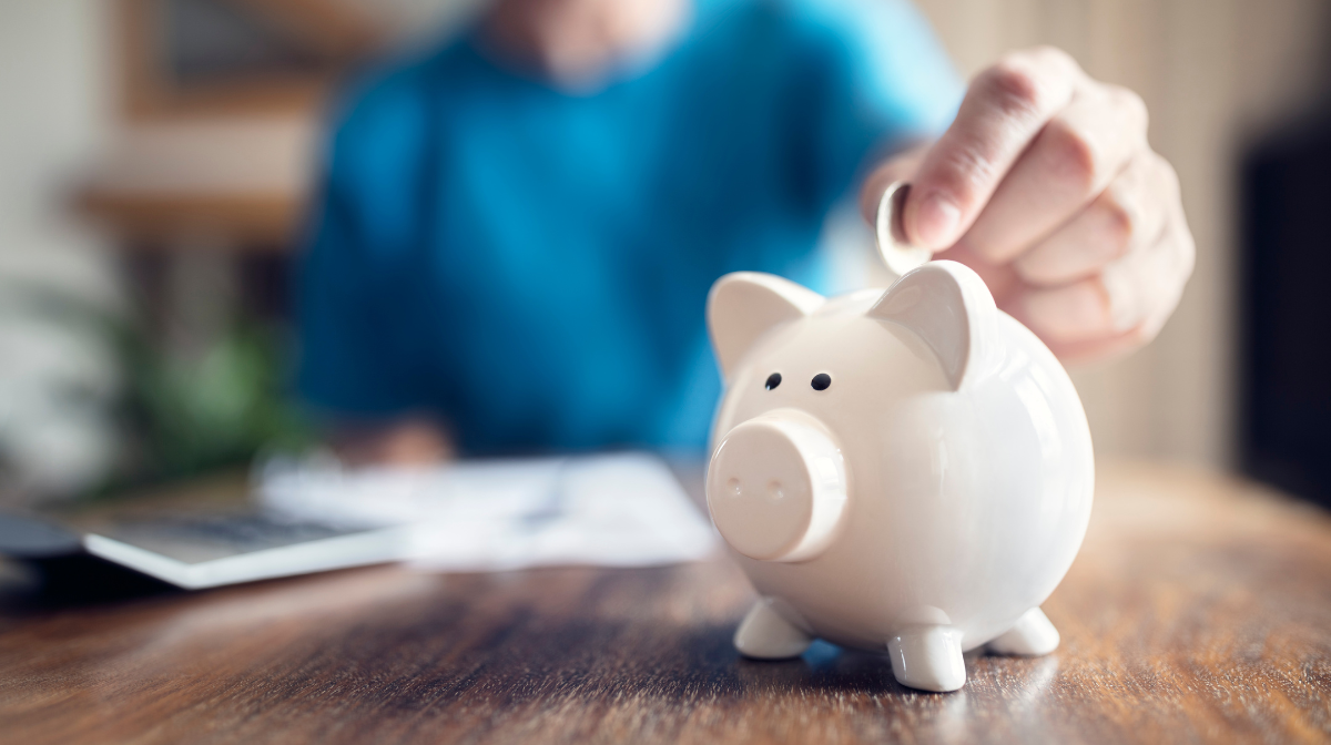 Person placing a coin into a white piggy bank on a wooden desk