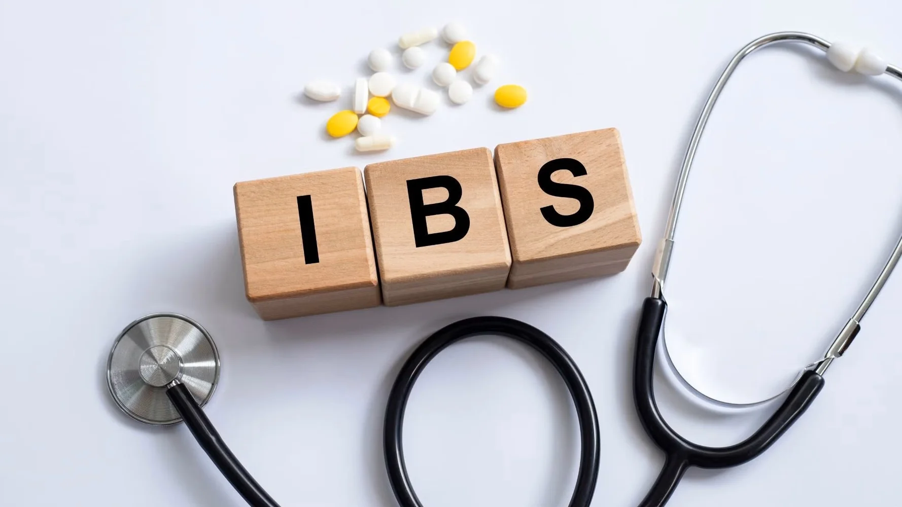 Wooden letter blocks spelling IBS next to a stethoscope and scattered pills, illustrating Irritable Bowel Syndrome