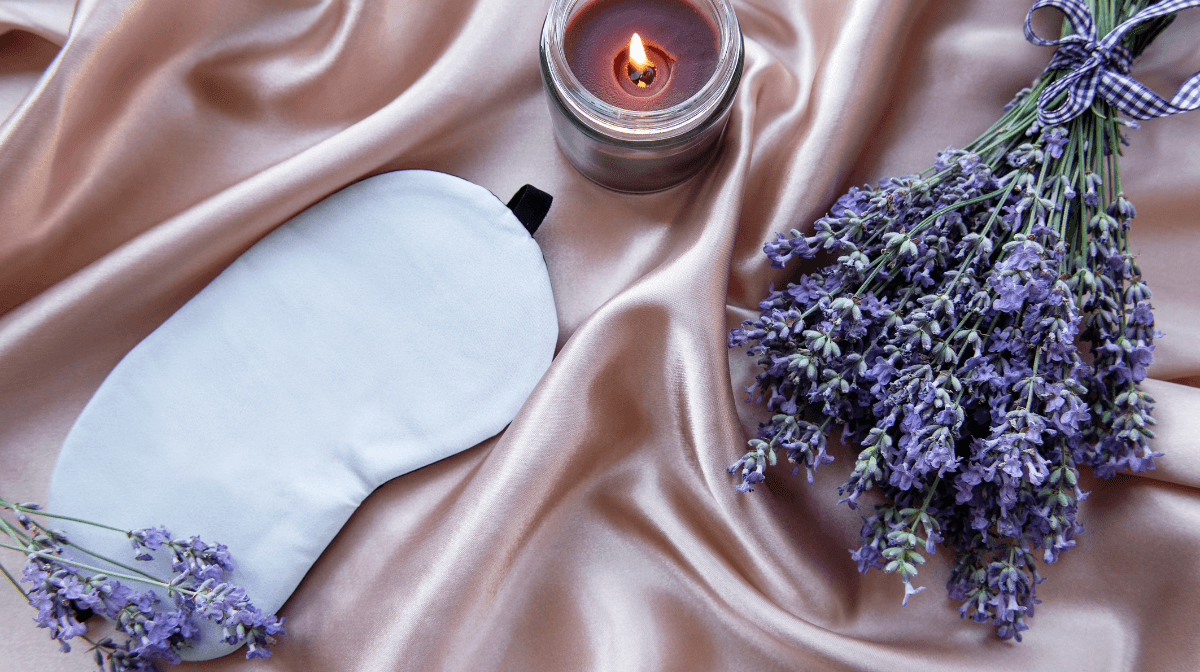 Sleep mask, lavender sprigs and a lit candle arranged on satin fabric, illustrating sleep improvement tips